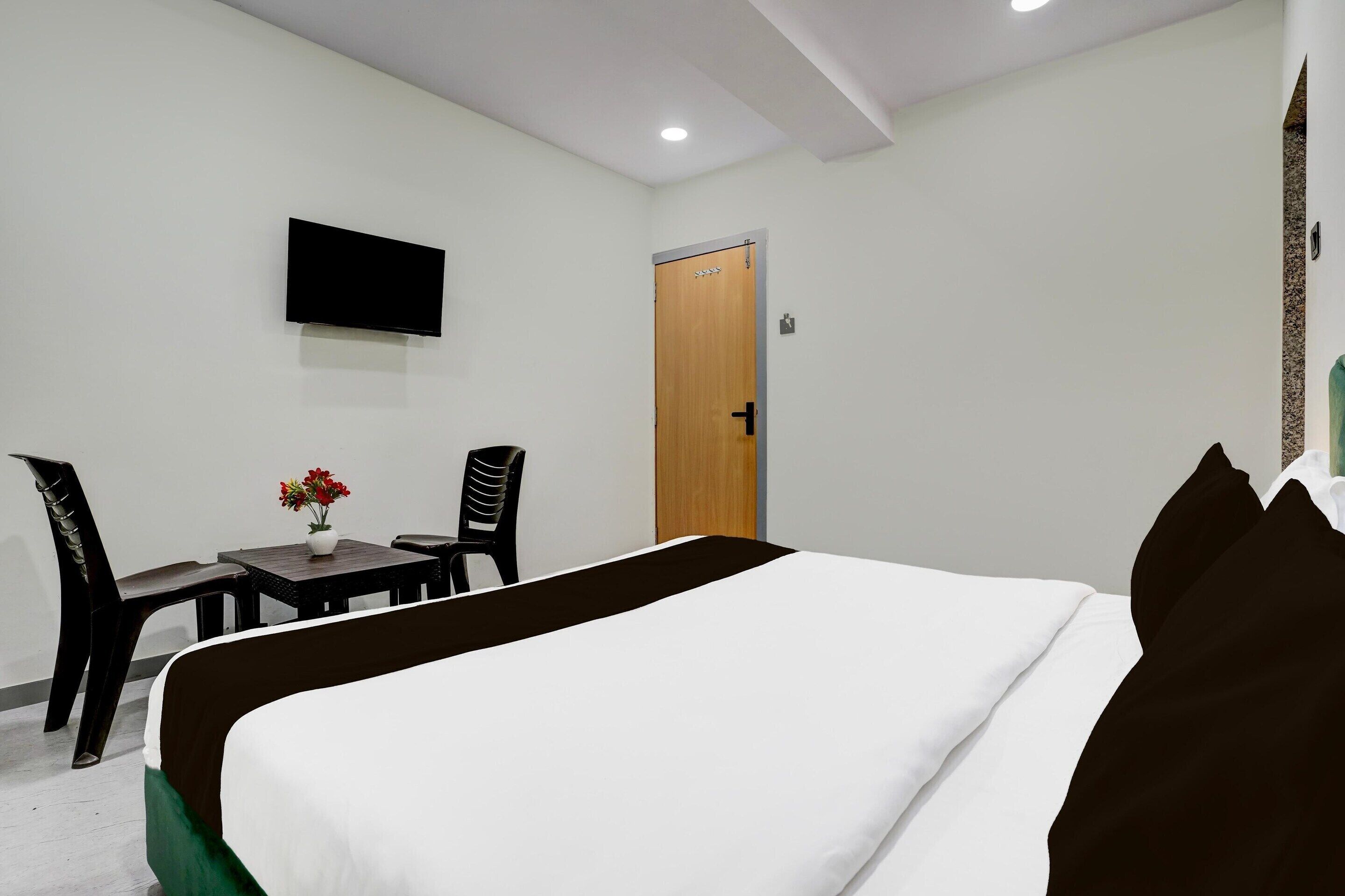 A hotel room with a bed, a TV mounted on the wall, a wooden door, and a small table with a vase of flowers.