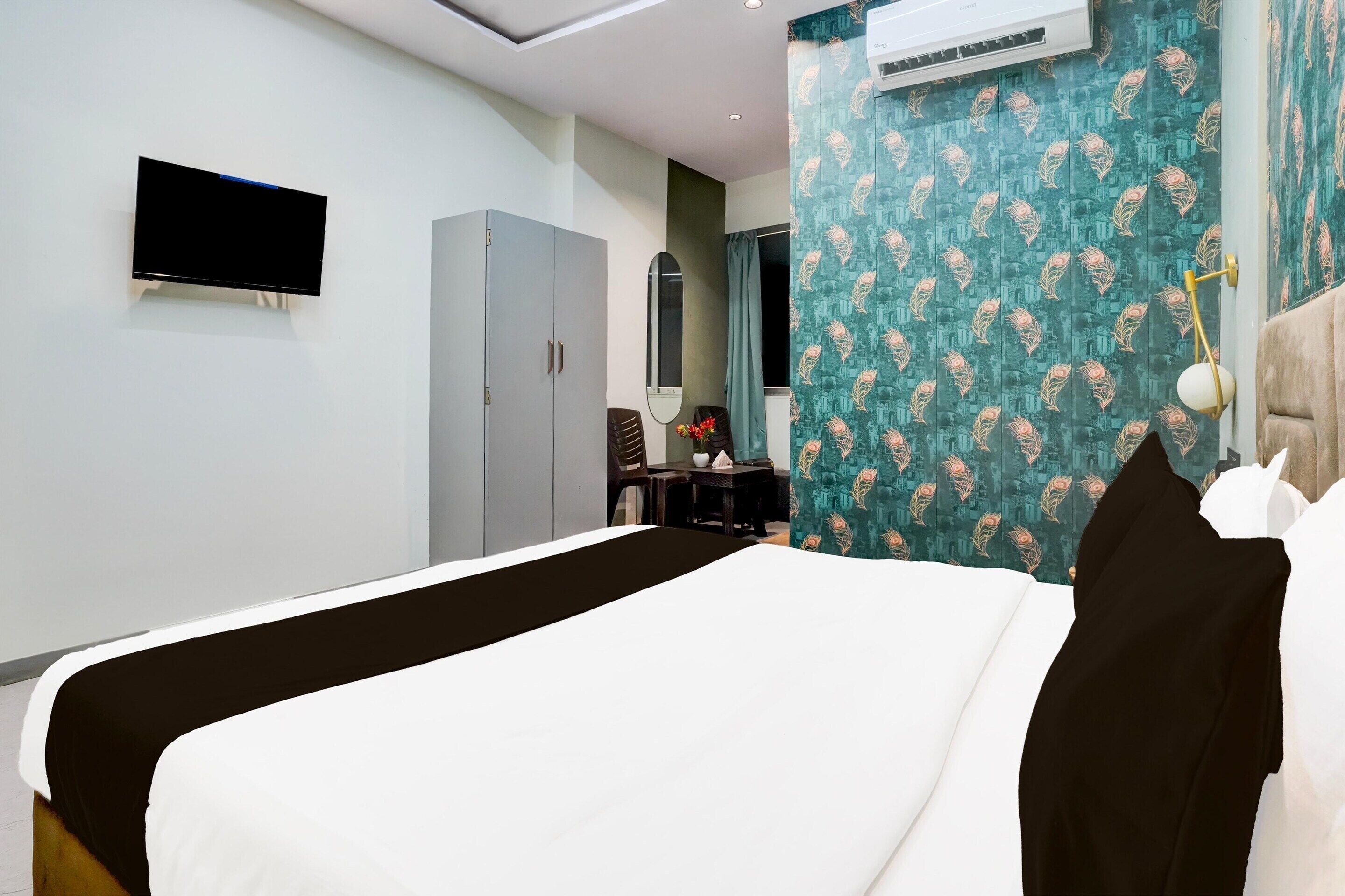 A hotel room with a bed, a flat-screen TV, a wardrobe, a desk with a chair, a mirror, and patterned curtains.