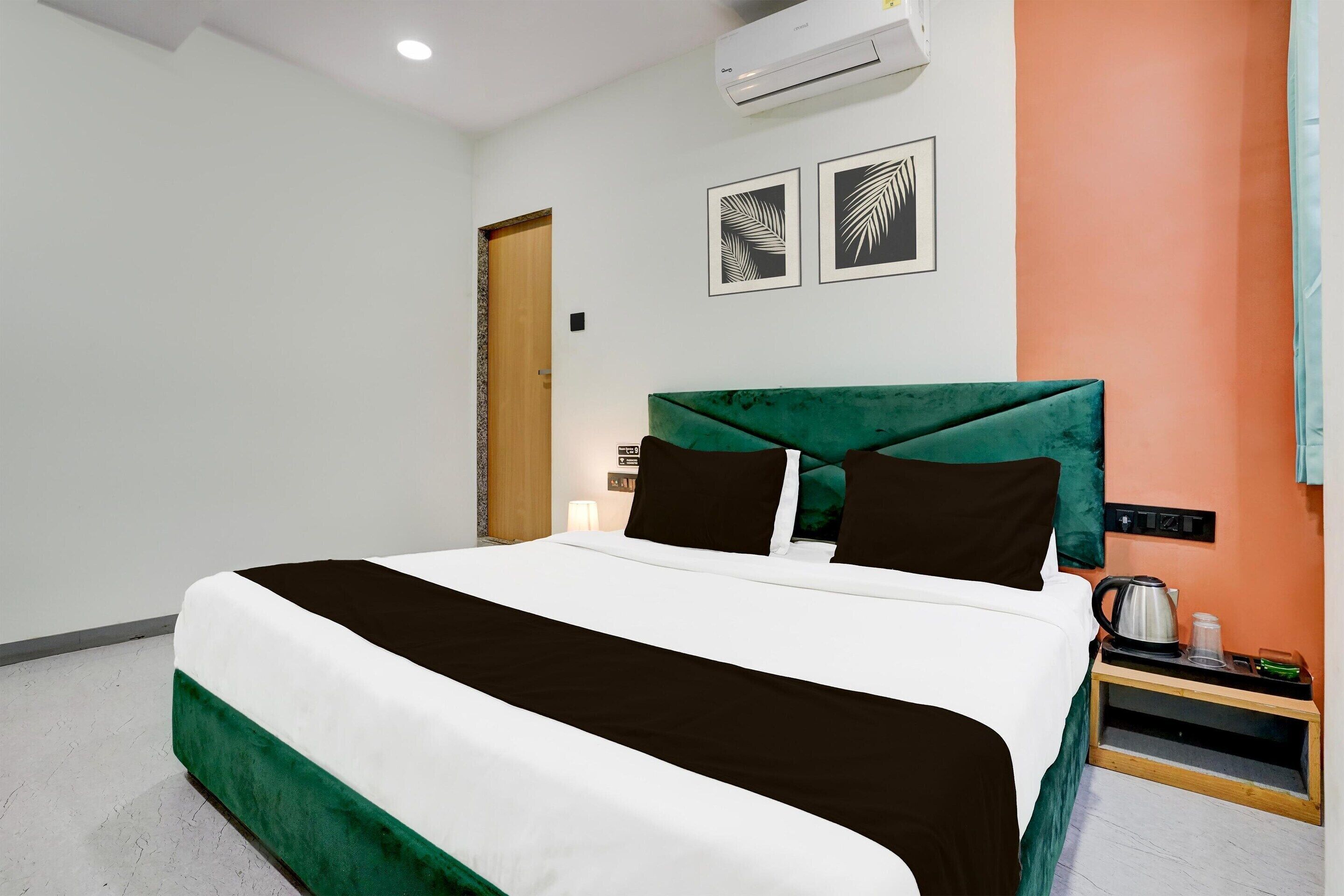 A neatly made bed with a green headboard, a bedside table with a kettle, and two framed leaf designs on the wall.