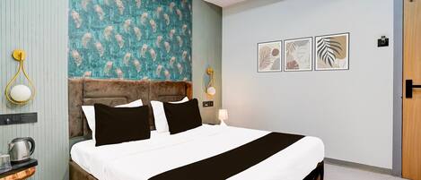 A modern hotel room with a large bed, a wooden headboard, a small side table with a kettle, and a door to the right.