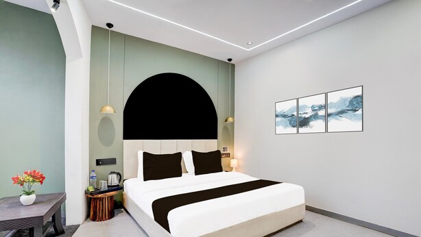 A modern hotel room with a large bed, a bedside table with a lamp, a small side table with a vase of flowers, and a wall-mounted artwork. - Deluxe Room Adajan Sky Palace (Surat)