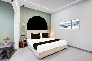 A modern hotel room with a large bed, a bedside table with a lamp, a small side table with a vase of flowers, and a wall-mounted artwork.