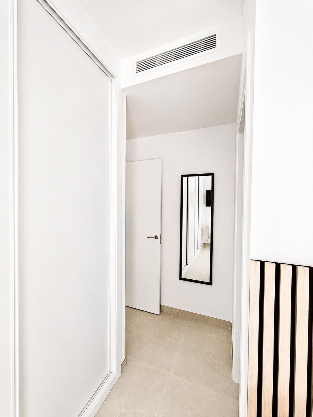 A hallway with a white door, a wall-mounted mirror, and a vent on the ceiling.