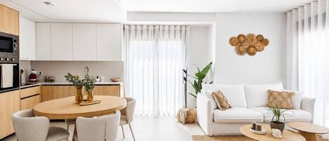 A modern kitchen with white cabinetry, a dining area with a round table and chairs, and a living room with a white sofa and a coffee table.