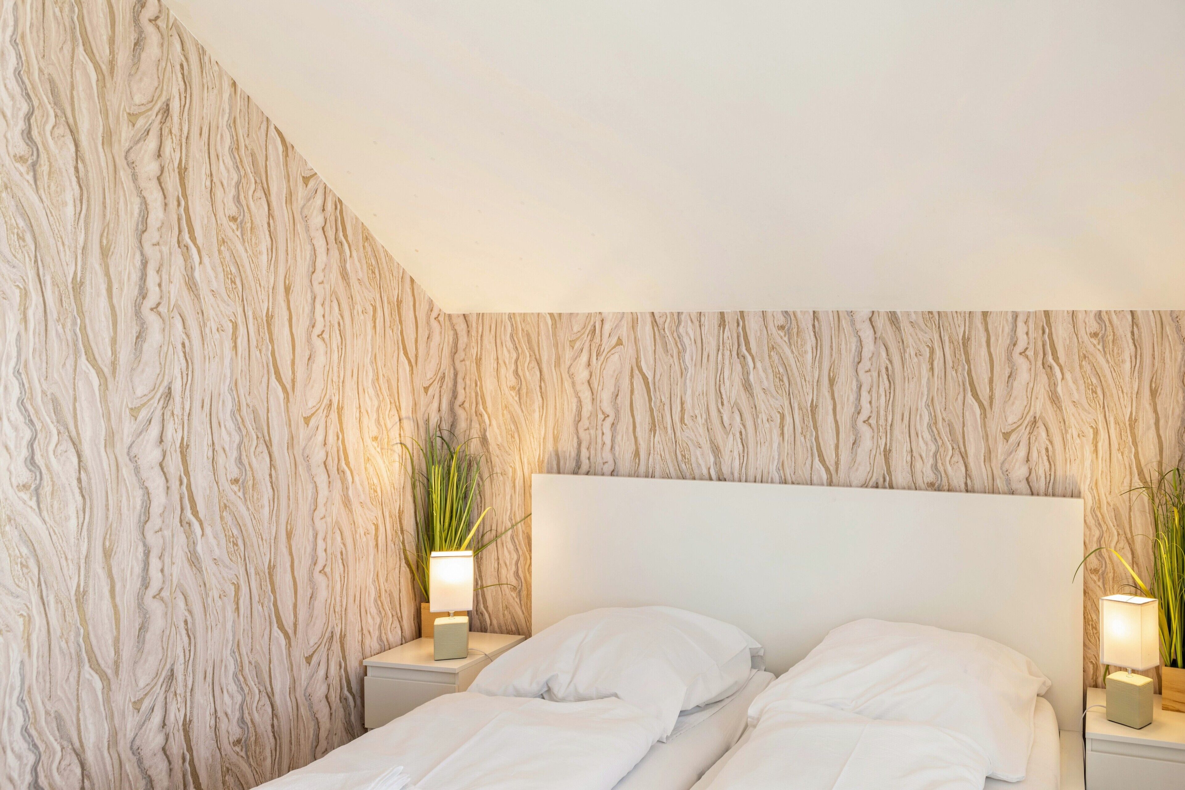 A bedroom with a marble-patterned wall, a white headboard, two bedside tables with lamps, and a bed with white bedding.