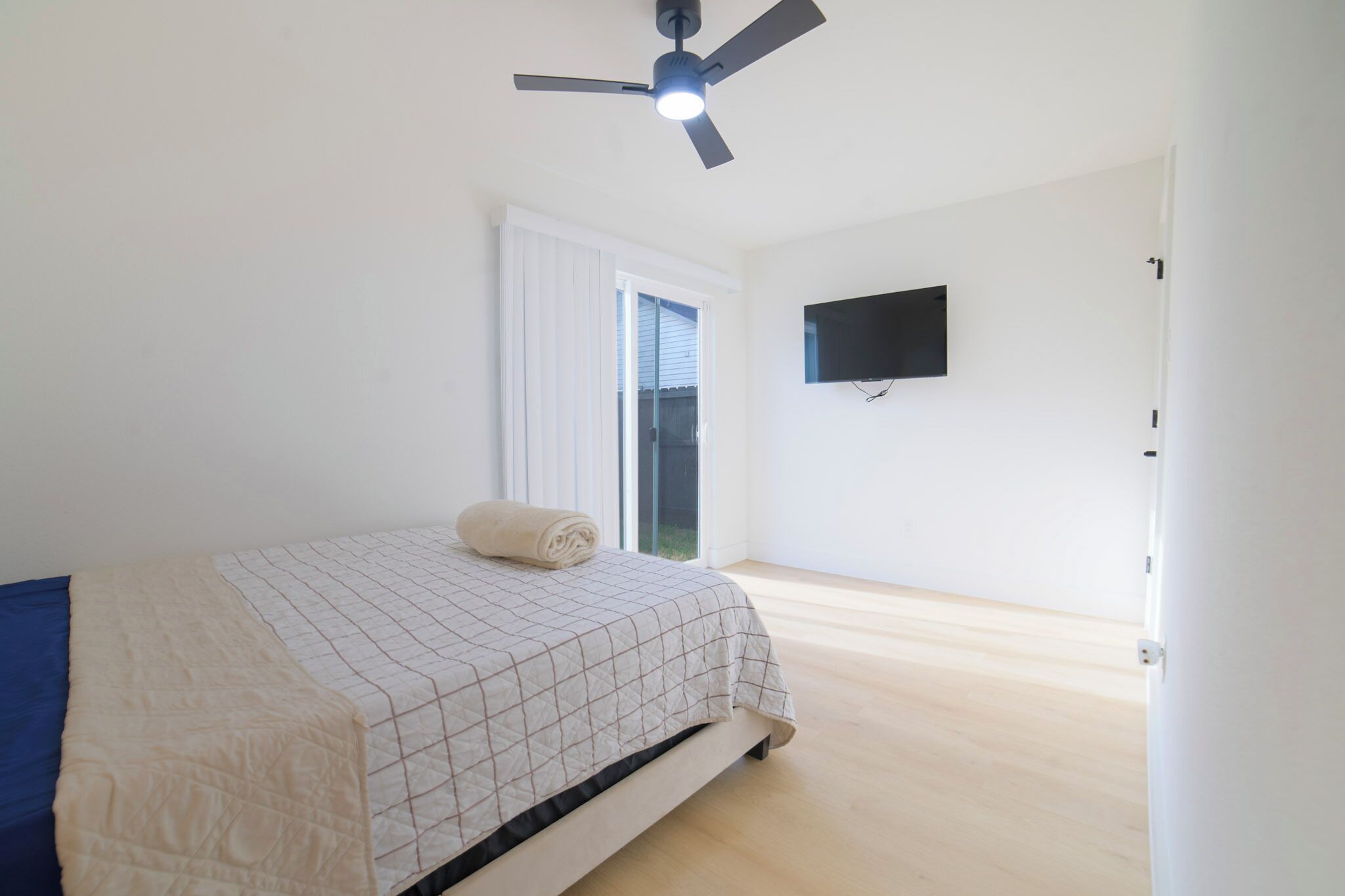 A bedroom with a bed, a ceiling fan, a TV mounted on the wall, and a sliding glass door.