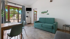 A room with a green sofa, a dining table with chairs, a TV mounted on the wall, and a view through large windows. - Holiday Home 'Kim' with Private Garden, Wi-Fi and Air Conditioning (Capoliveri)