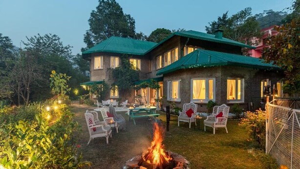 A two-story building with a green roof, outdoor seating area with white chairs, and a fire pit in the center. - Alaya Stays Chalet Vie 3 BHK Villa (Kasauli)