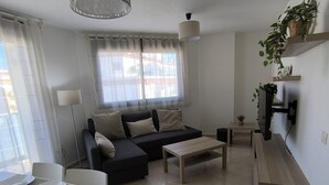 Een moderne woonkamer met een bank, salontafel, eettafel en stoelen. Er is een televisie aan de muur bevestigd en een raam met gordijnen.