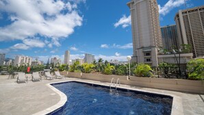 Una piscina en la azotea con sillones de descanso, rodeada de palmeras y edificios.