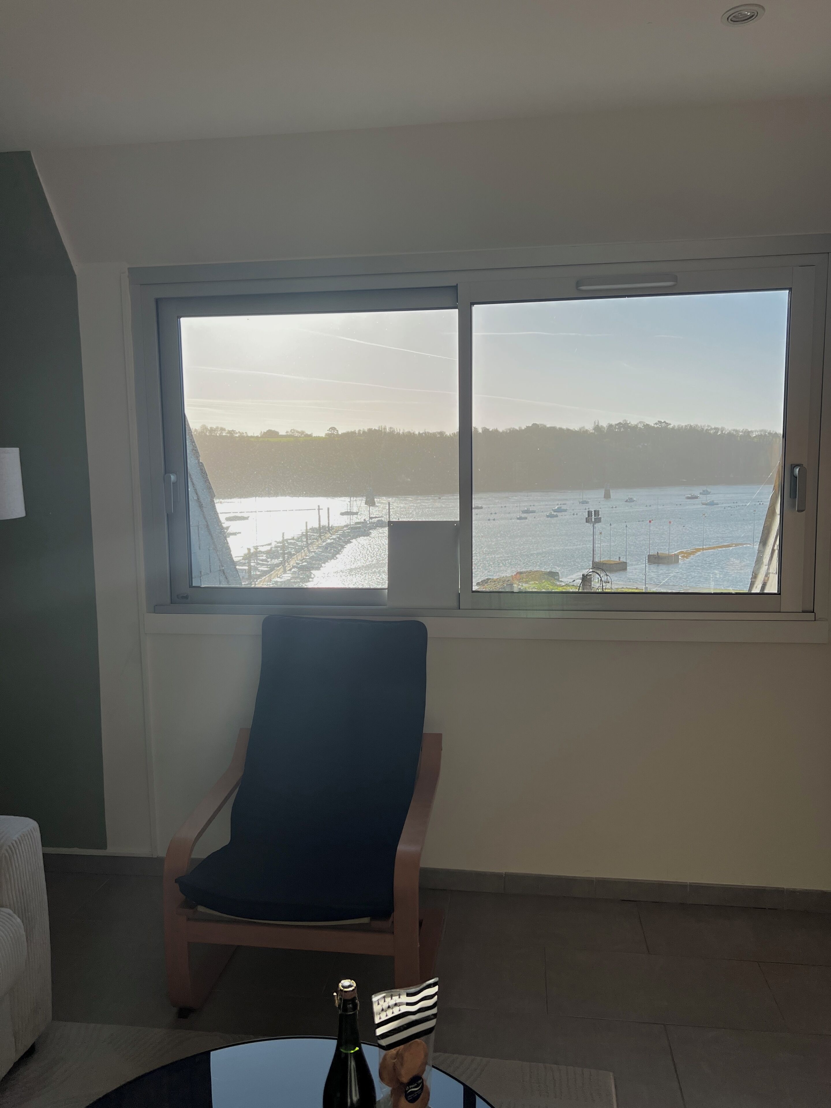 A room with a large window, a chair, a small table with a bottle, and a teddy bear.