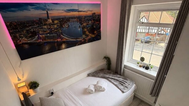 A small bedroom with a bed, a window with curtains, a wall-mounted painting, and a bedside table with a lamp. - 4Bed House with parking and GYM /London/Edgware (Edgware)