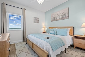Ein Schlafzimmer mit Bett, Nachttisch, Vorhängen am Fenster, Deckenventilator und Kunstwerk an der Wand.