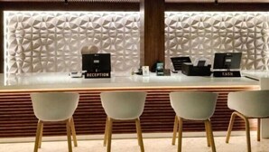 A reception desk with a modern geometric wall design, a cash register, a reception sign, and three white chairs in front. - Hotel O Chil SEZ Coimbatore (Coimbatore)