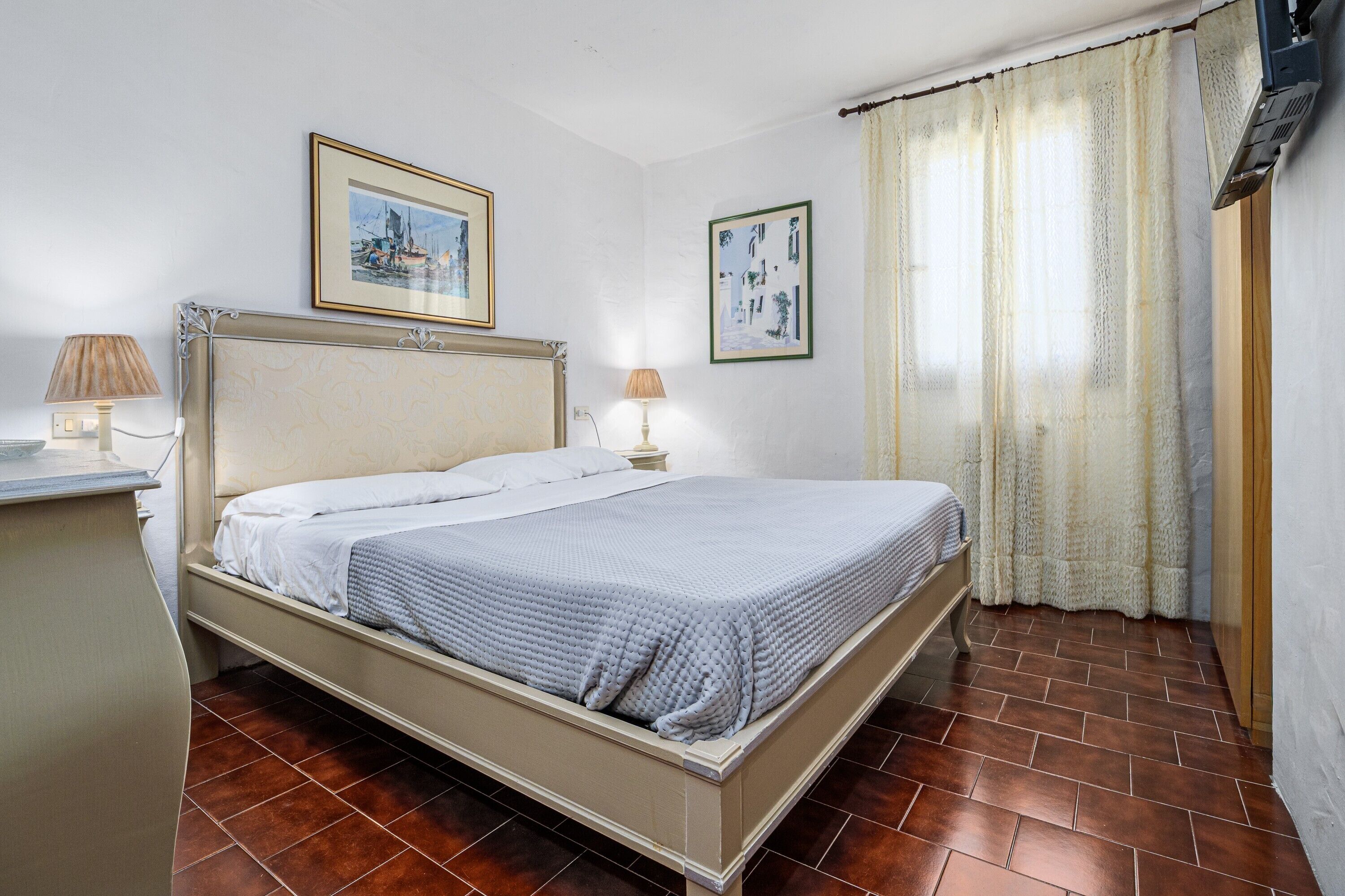 A bedroom with a bed, two bedside lamps, a framed picture above the bed, a window with curtains, and a radiator.