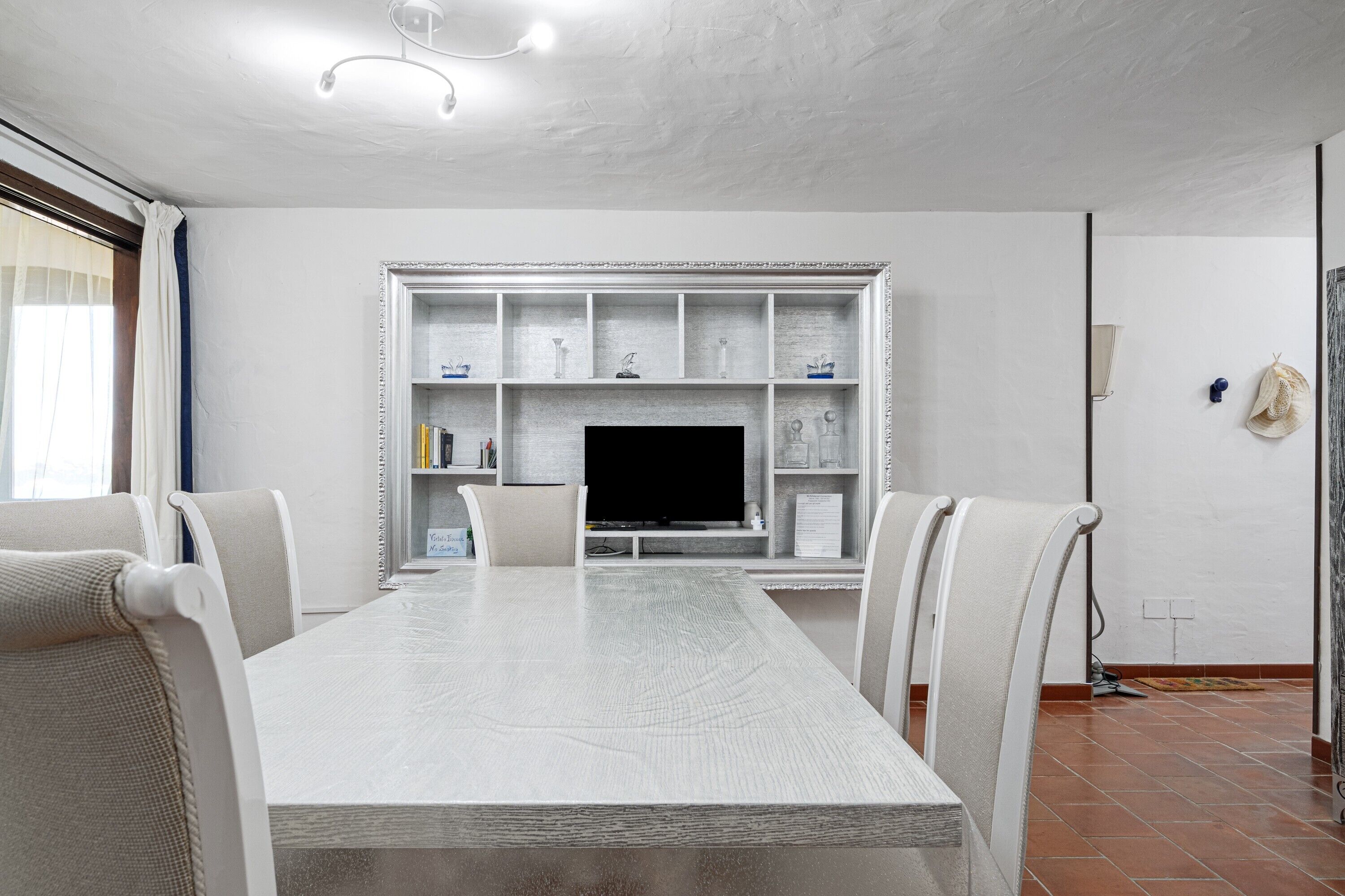 A dining area with a large table, chairs, a television, and a shelf with decorative items.
