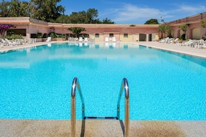 Una piscina cristallina con una scala metallica che vi si immerge, circondata da lettini e un edificio con archi e tetto piastrellato.