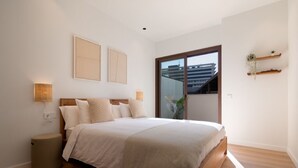 Une chambre à coucher comprenant un lit, des lampes de chevet, un petit tabouret et une porte-fenêtre coulissante donnant sur un balcon.