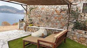 Un espace extérieur aménagé avec des murs en pierre, offrant une vue sur la mer, agrémenté d’une pergola et d’une petite table avec une chaise.