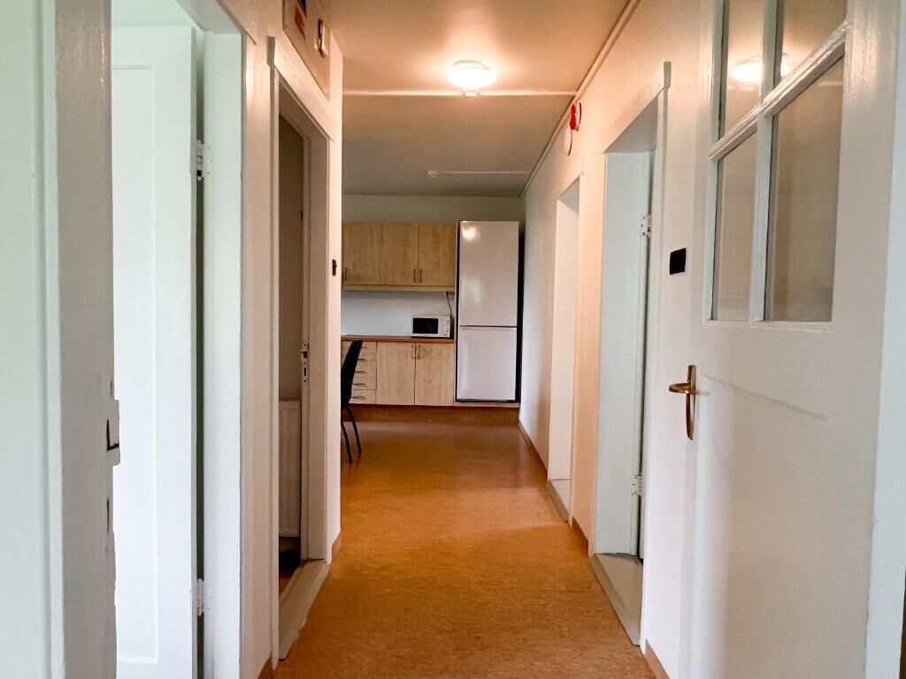 A narrow hallway with white doors, a kitchen area with cabinets and appliances, and a carpeted floor.