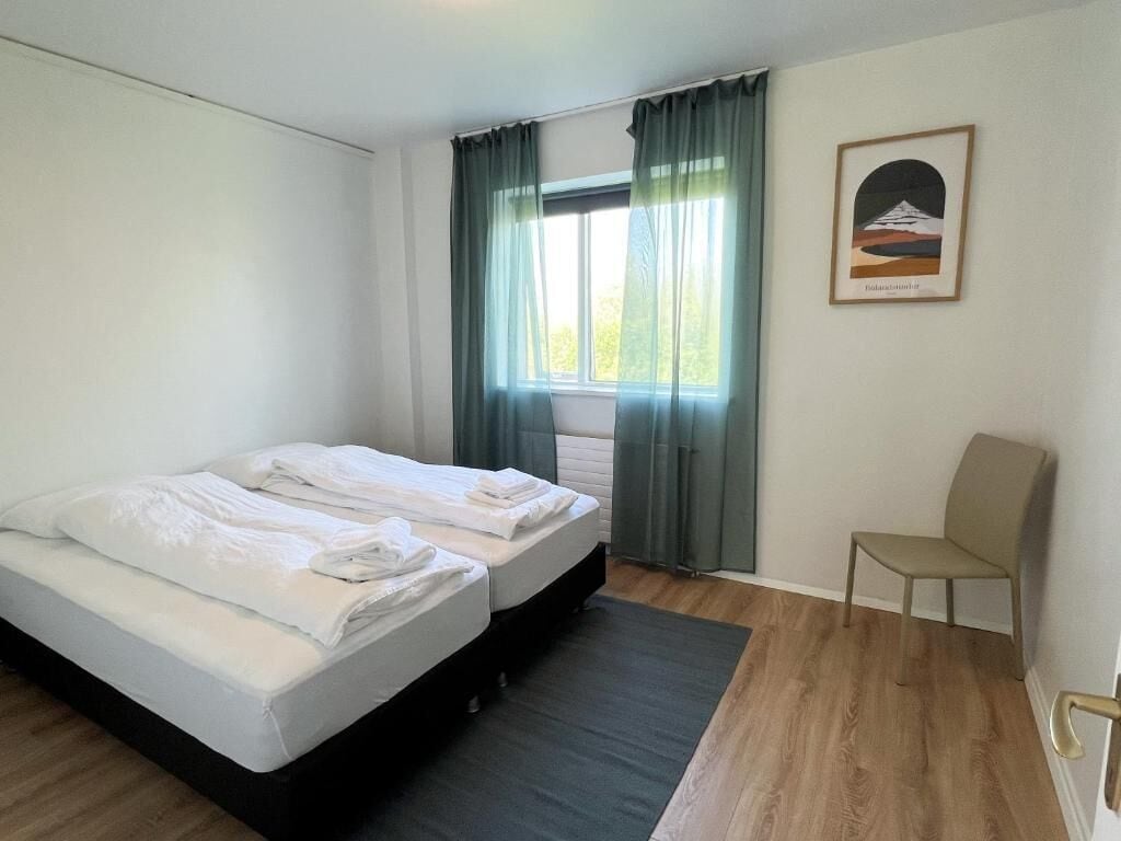 Una habitación con dos camas, una silla, un ventanal con cortinas y un cuadro en la pared.