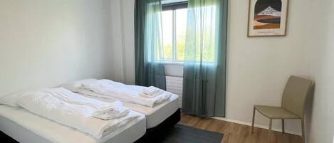 Una habitación con dos camas, una silla, un ventanal con cortinas y un cuadro en la pared.