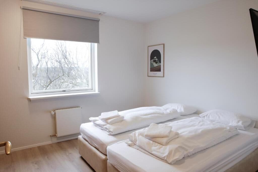 Una habitación con dos camas, un radiador, un ventanal con vistas a árboles y un cuadro en la pared.