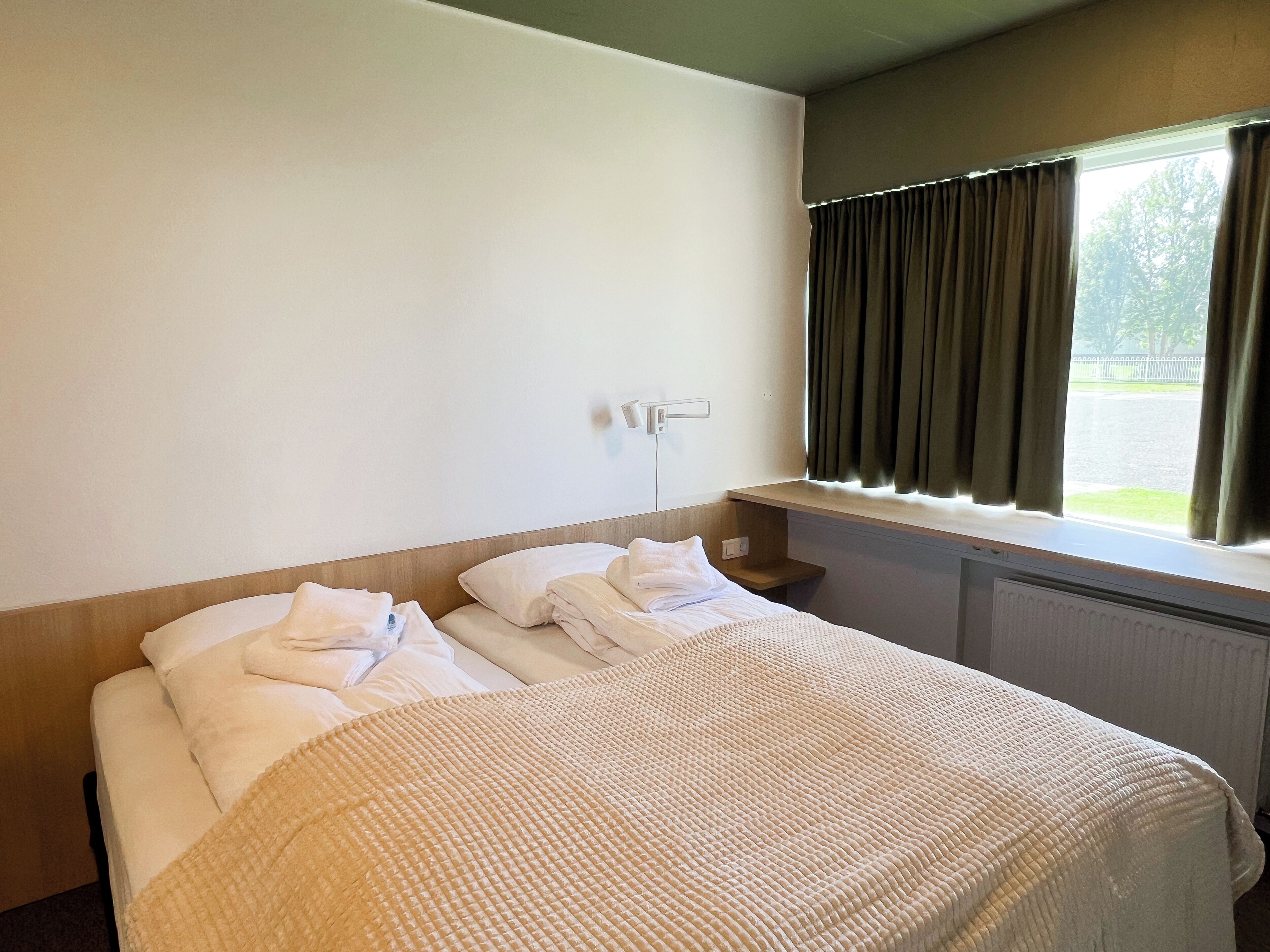 A hotel room with a bed, a nightstand, a window with curtains, and a view of the outside.
