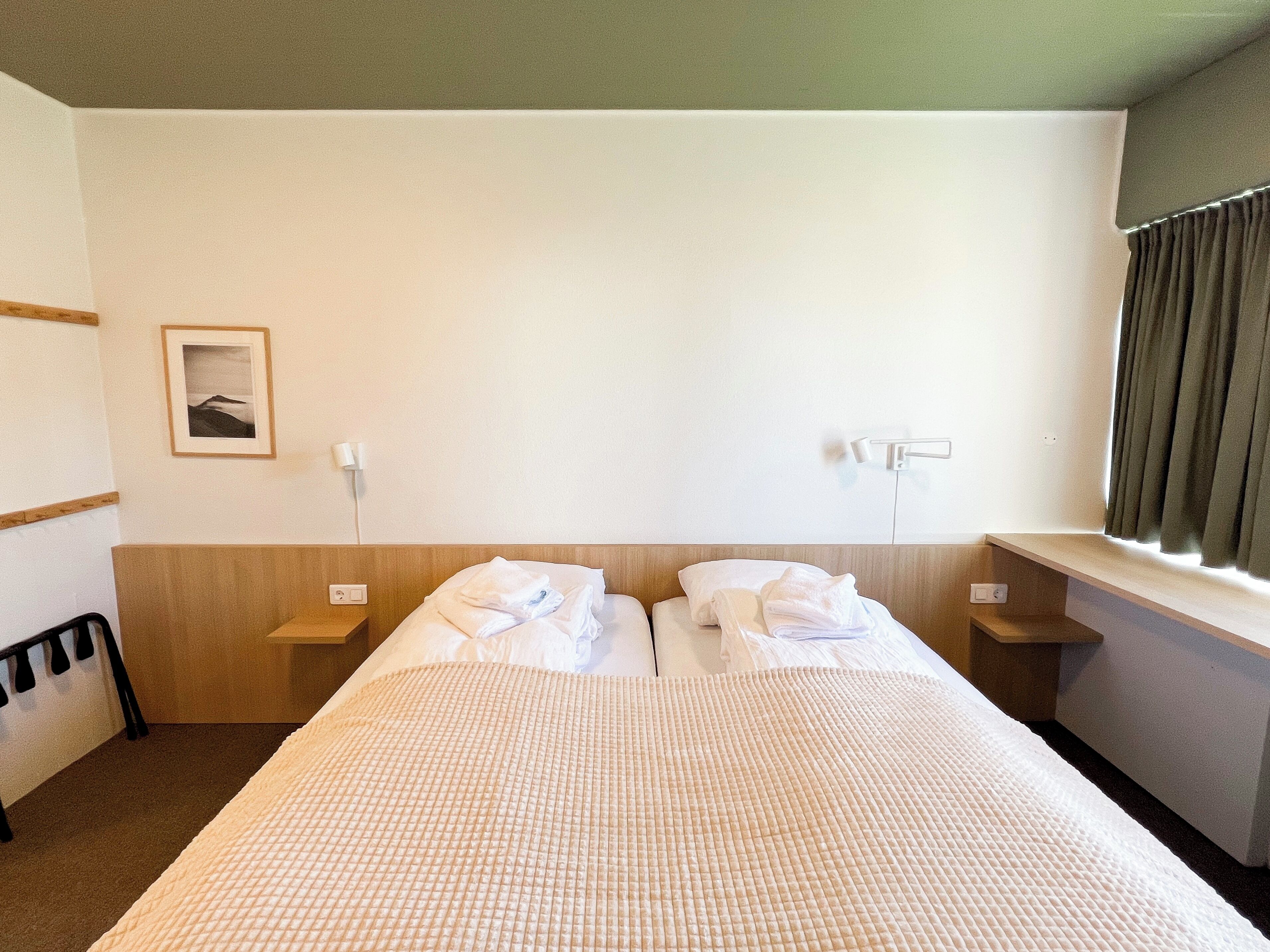 Habitación de hotel con cama, dos mesitas de noche, una lámpara de pared, un cuadro enmarcado y una ventana con cortinas.