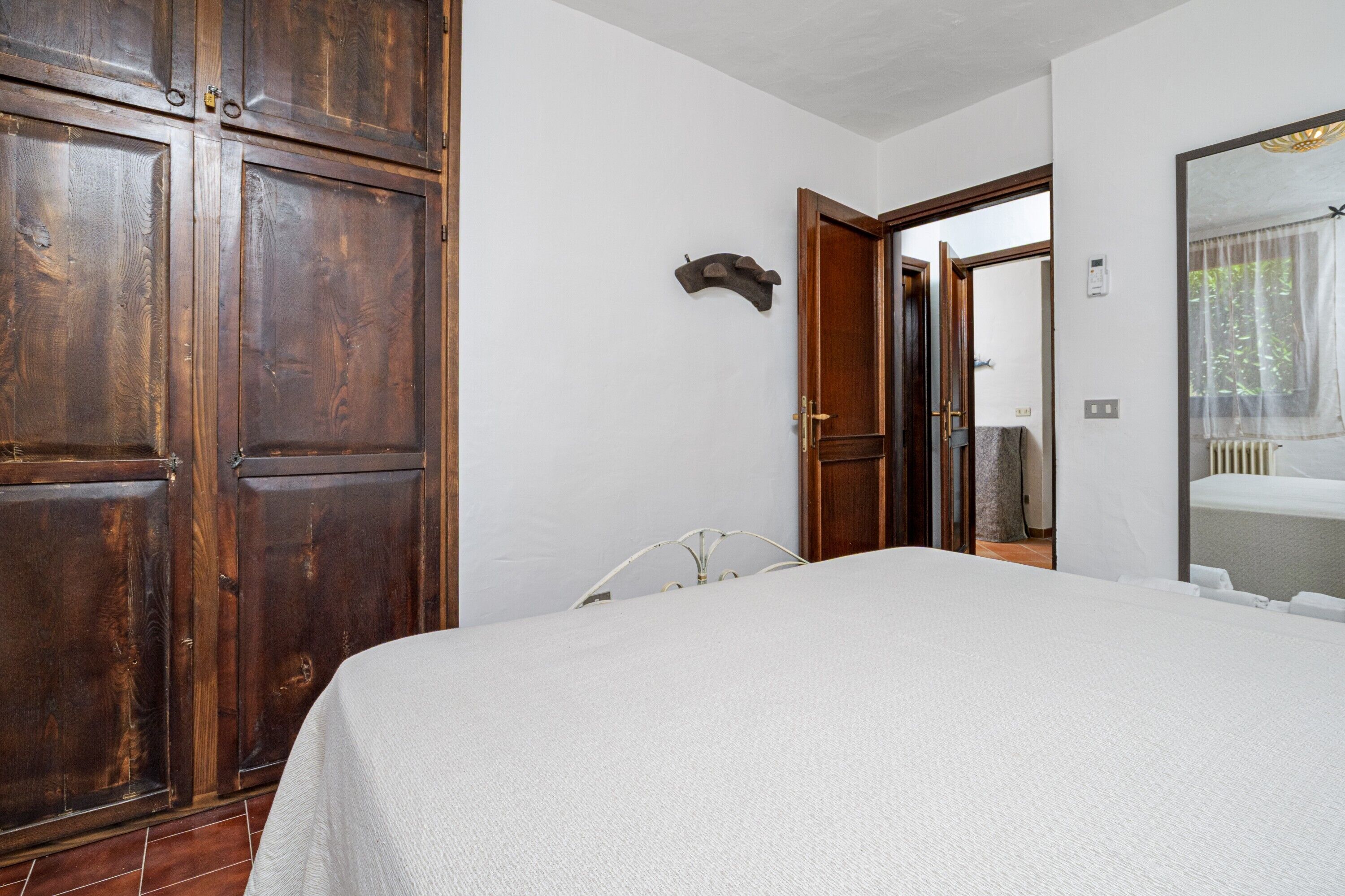 A bedroom with a large bed, wooden wardrobe, and a mirror reflecting a window.