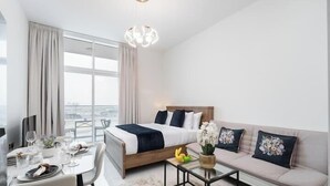 A hotel room with a bed, a sofa, a dining table with chairs, and a view through large windows. - Vogue | Skyz by Danube near Dubai Miracle Garden (Dubai)