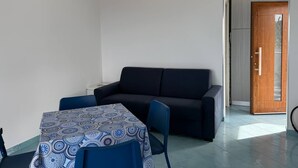 Een kleine, moderne kamer met een bank, een tafel met een tafelkleed met patroon, en een deur naar een buitengebied.