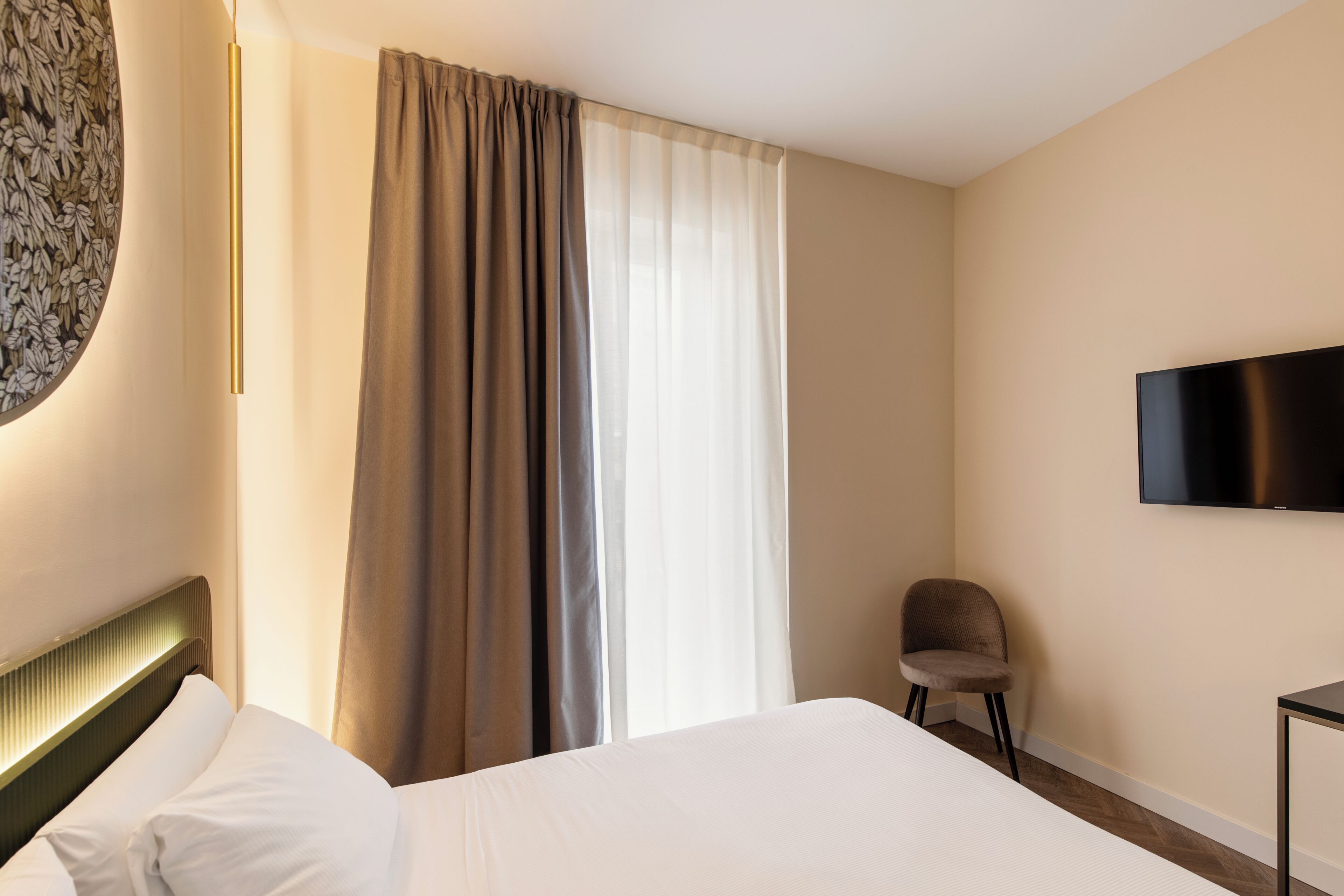 Une chambre d’hôtel avec un lit, une chaise, une télévision et une fenêtre avec des rideaux.
