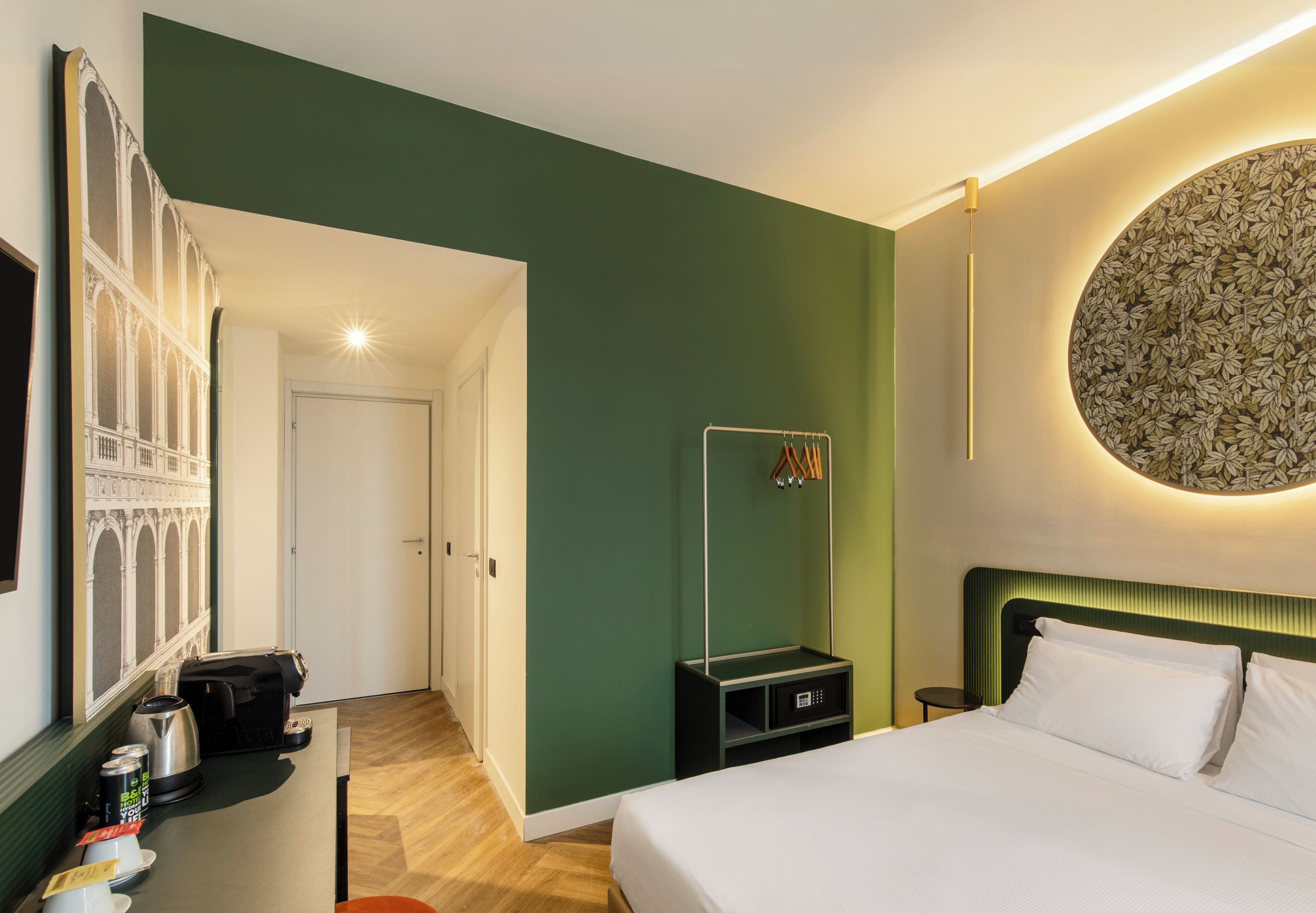 A hotel room with a bed, a desk with a kettle, a small shelf, and a wall-mounted circular artwork.