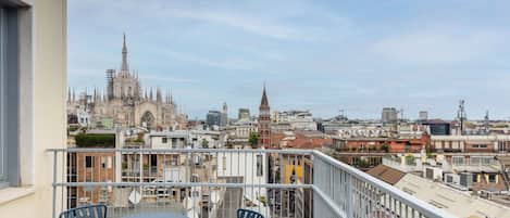 Un balcón con dos sillas y una mesa, con vistas a un paisaje urbano que incluye edificios históricos y construcciones modernas.