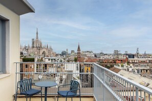 Un balcón con dos sillas y una mesa, con vistas a un paisaje urbano que incluye edificios históricos y construcciones modernas.