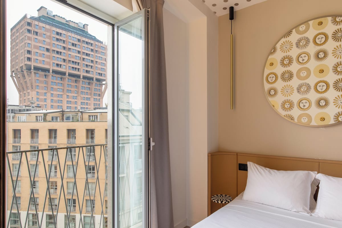 Habitación de hotel con cama, un adorno circular en la pared, ventana con cortina y vista a un edificio.
