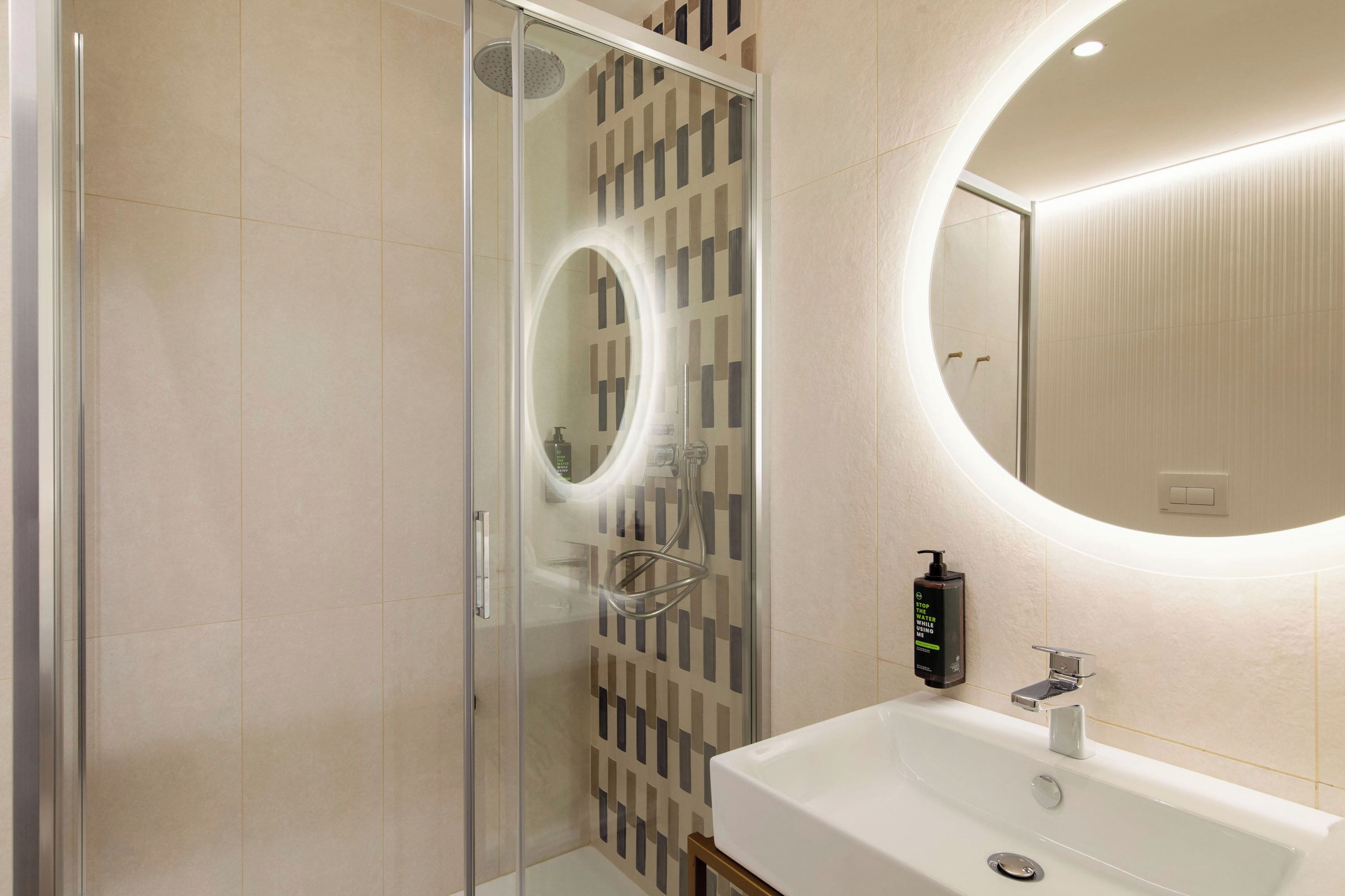 A modern bathroom with a glass-enclosed shower, a round mirror, and a white sink.