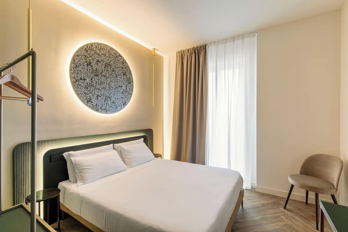 Habitación de hotel con cama, un adorno redondo en la pared, una silla y una ventana con cortinas.