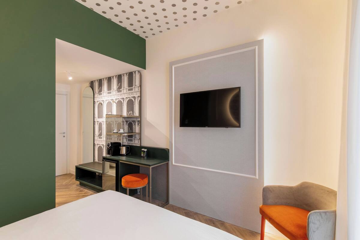 Una habitación de hotel moderna con cama, escritorio y silla, televisión montada en la pared y vista a un edificio a través de la ventana.