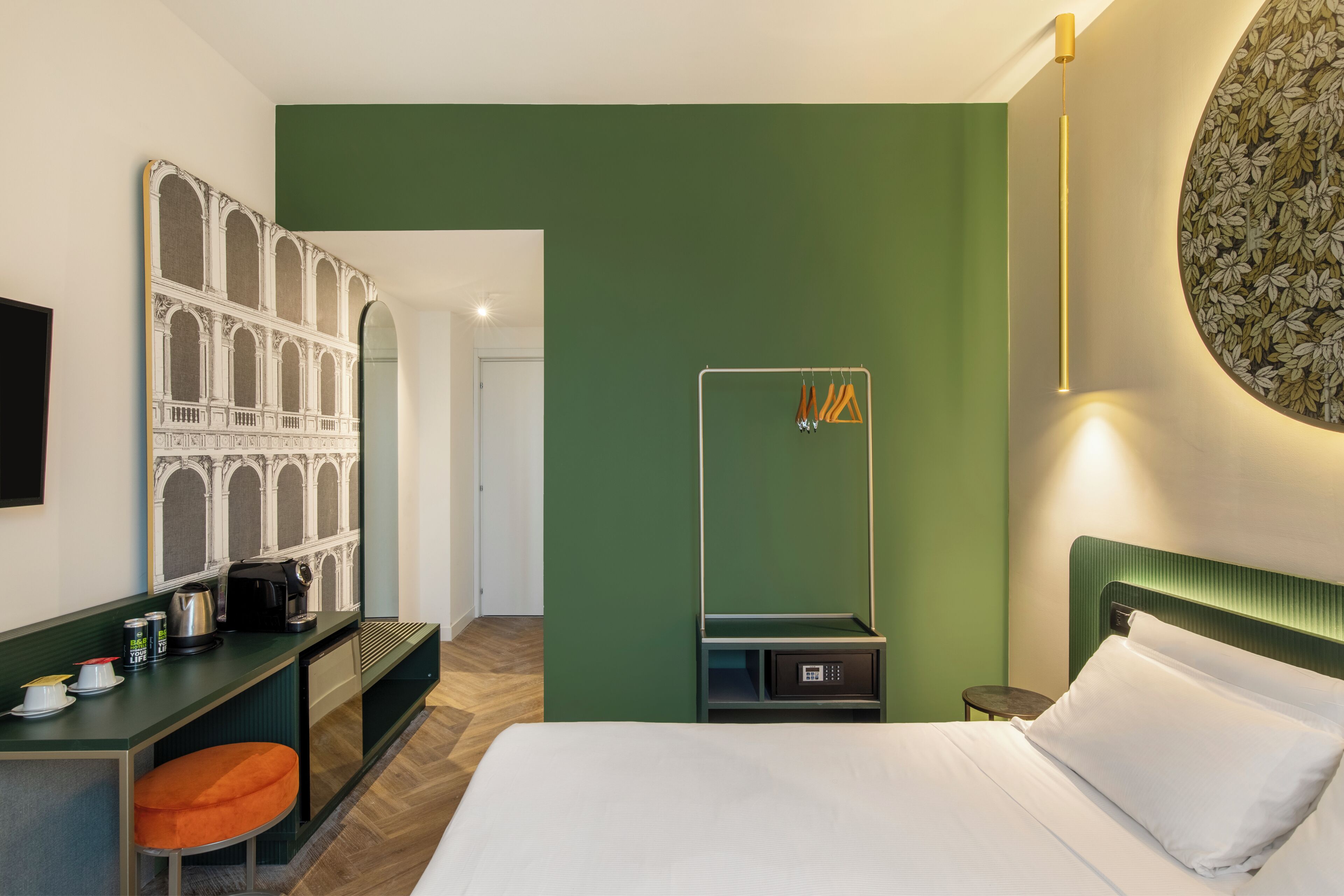 A hotel room with a bed, a desk with a kettle, a small table, and a hanging clothes rack.