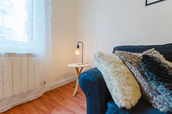 Ett rum med ett fönster, ett litet bord med en lampa, en soffa med en fäll av fuskpäls och ett element.