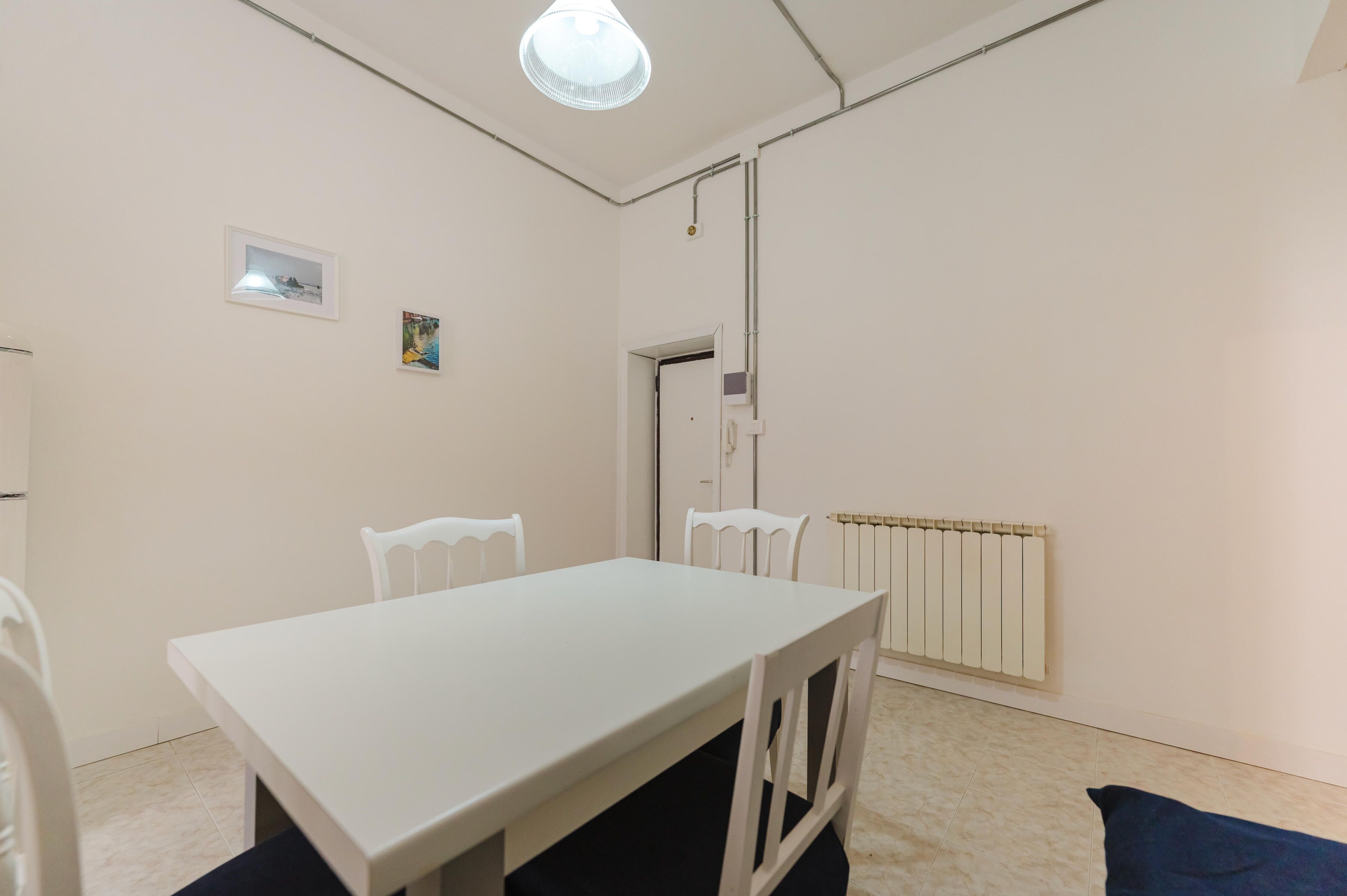 Una habitación con una mesa blanca, cuatro sillas, una lámpara en el techo, un radiador y dos cuadros en la pared.