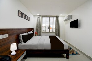 Une chambre à coucher avec une tête de lit en bois, un lit recouvert d’une couette blanche et marron, une lampe de chevet, un téléviseur à écran plat fixé au mur et une fenêtre avec des rideaux.