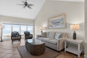 Ein Wohnzimmer mit Blick auf den Strand, einem Deckenventilator, einem Sofa, zwei Stühlen, einem Couchtisch und zwei Lampen.
