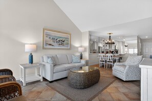 Ein modernes Wohnzimmer mit einer Couch, einem Couchtisch und Sesseln, ein Essbereich mit Tisch und Stühlen sowie eine Küche im Hintergrund.