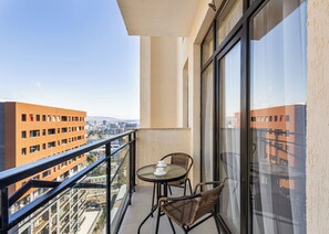 Un balcón con una mesa de vidrio y dos sillas de mimbre, con vista a un paisaje urbano con edificios modernos.