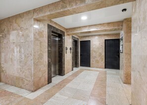 Um corredor com piso de mármore, dois elevadores e portas escuras.