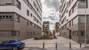 Een moderne stadstraat met meerlaagsgebouwen, een blauwe auto geparkeerd aan de kant, en een heldere lucht.