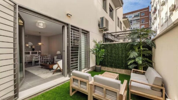 Een modern appartement met een schuifdeur naar een balkon, een klein terras met een bank en tafel, en een groene wand vol planten.
